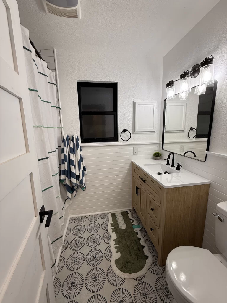 A modern bathroom featuring a stylish wooden vanity with a white sink, a large mirror, and black fixtures. A shower curtain with green and white stripes is visible, along with a blue-striped towel and a decorative green rug shaped like an alligator. The floor has a circular patterned design and there is a window providing natural light.