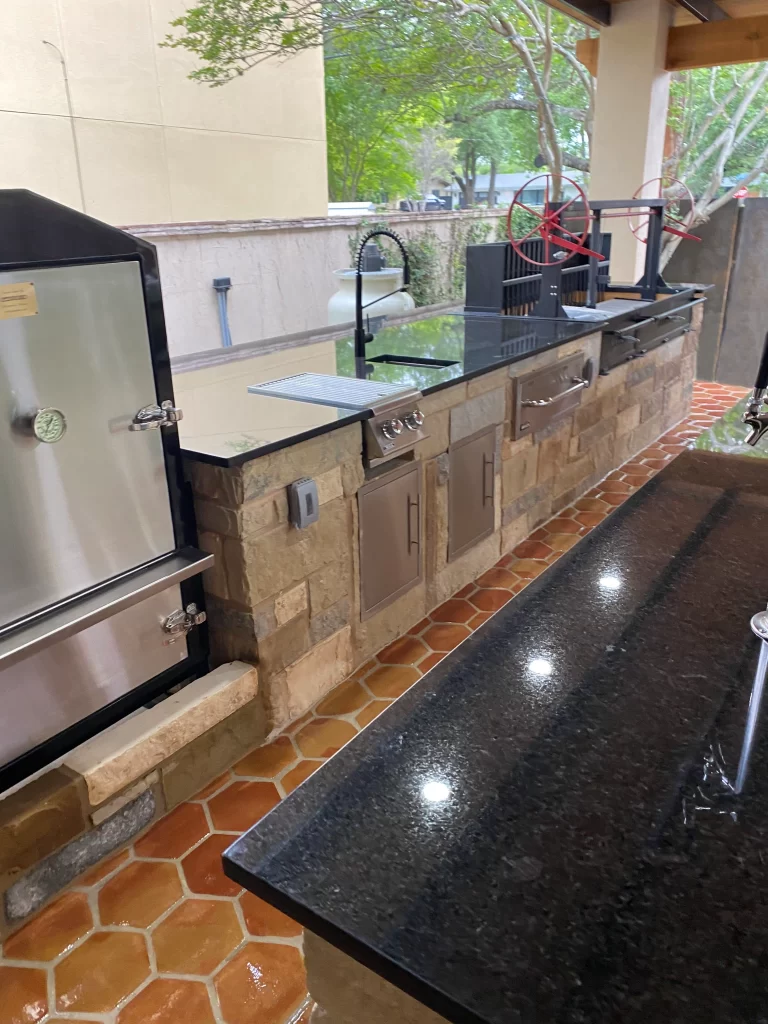 An outdoor kitchen featuring a granite countertop, stainless steel appliances, and a stone base. The setup includes a grill, storage cabinets, and a faucet, surrounded by a tiled floor and greenery in the background.