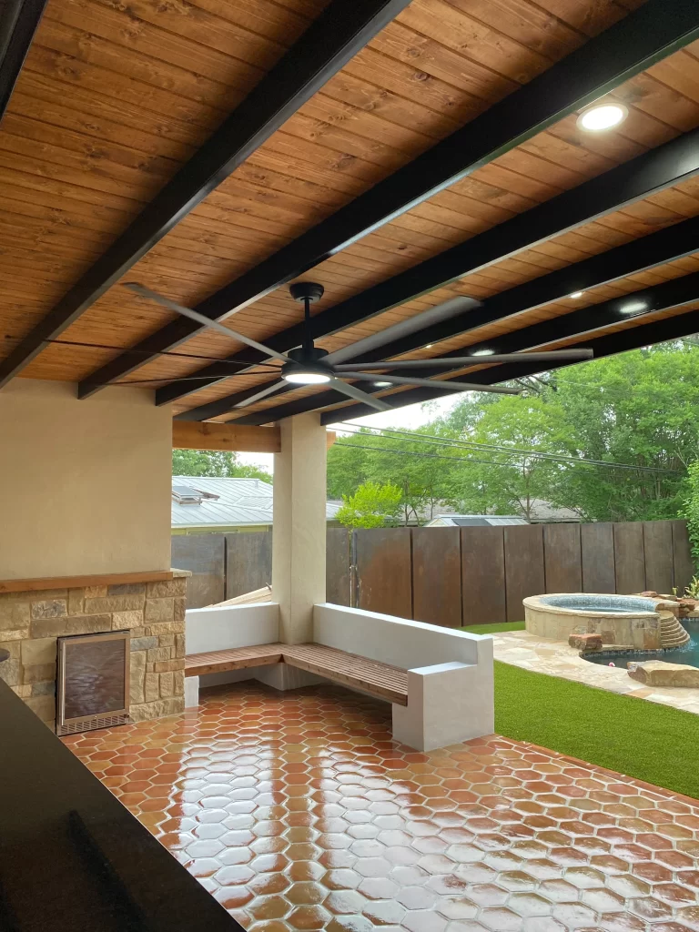 Covered outdoor patio with wooden ceiling, ceiling fan, and built-in stone fireplace, featuring a tiled floor and a garden view.