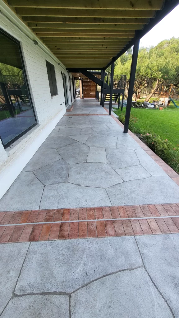 A view of a patio with a decorative concrete floor featuring stone patterns and brick accents, leading to a green lawn and wooden structures.