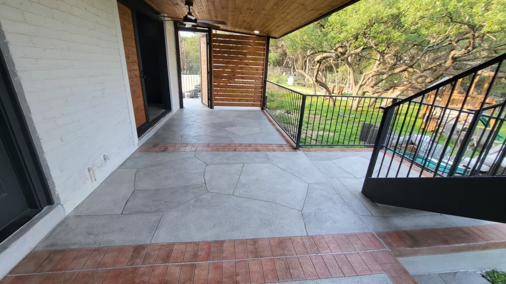 A modern outdoor patio area with textured concrete flooring and a wooden ceiling. The patio has a railing and a view of a green garden, visible through an open door.