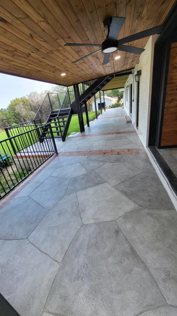 An outdoor patio area with a textured stone floor, wooden ceiling, and a ceiling fan, featuring stairs in the background leading to a grassy area.